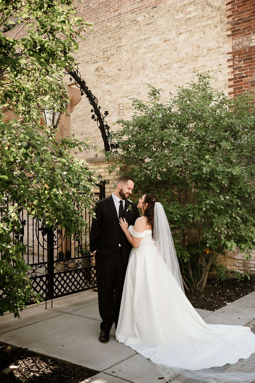 Bride and groom stand together in a garden courtyard, gazing at each other with smiles, framed by iron gates and greenery.