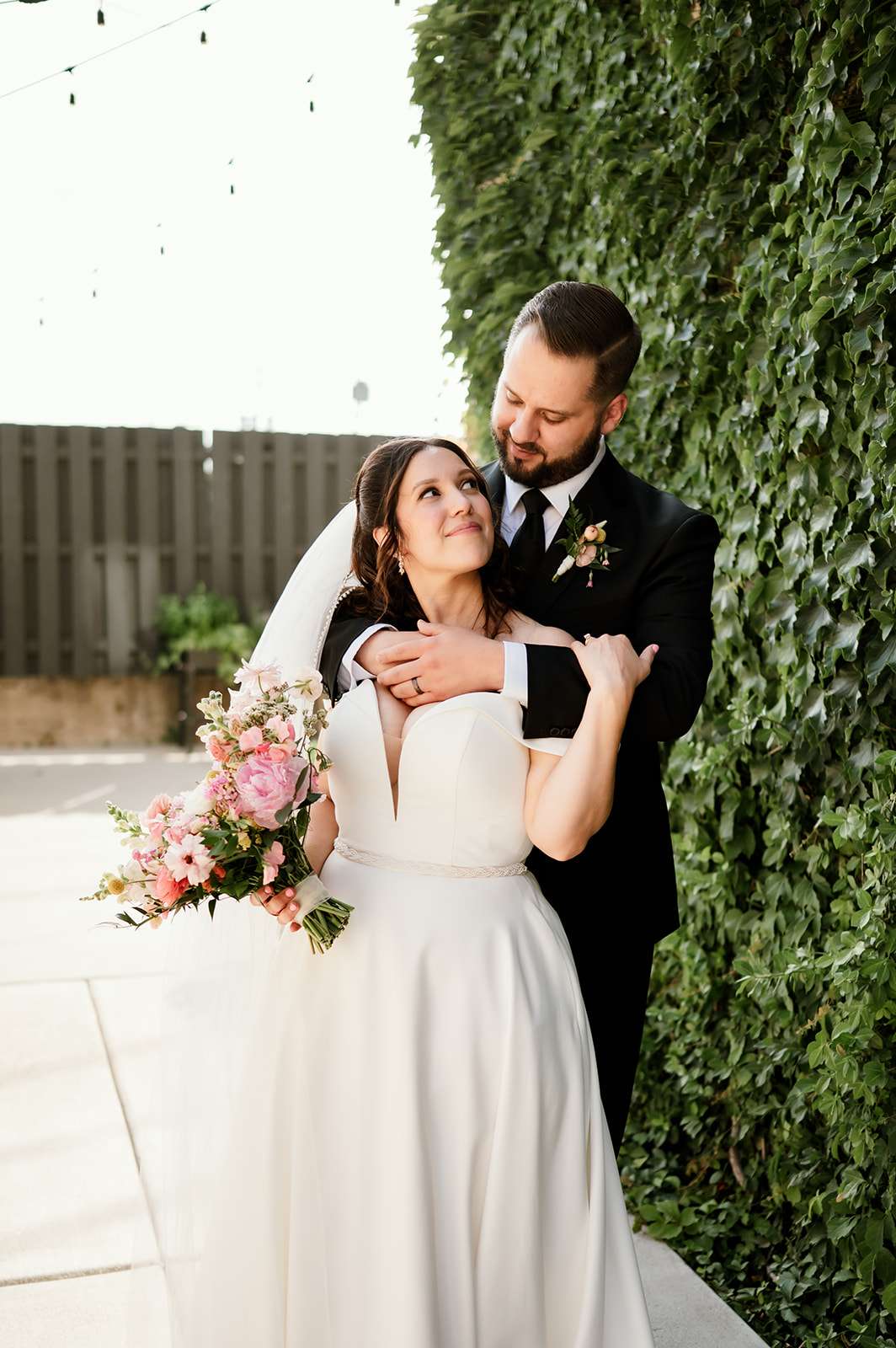 Bride and groom embrace affectionately in front of an ivy-covered wall, with the bride holding a bouquet of pink flowers and smiling up at the groom.