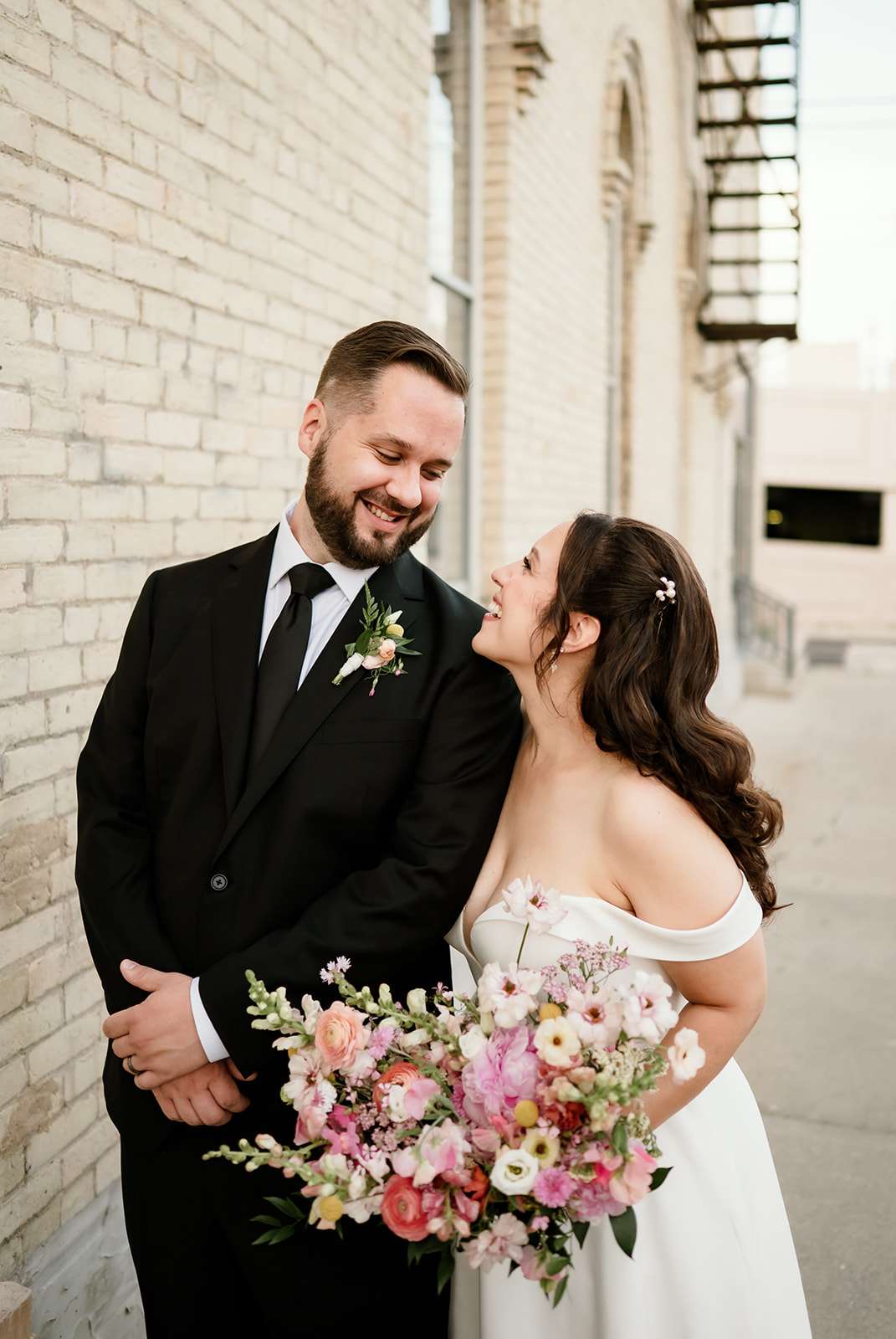 Bride and groom smile at each other in front of a cream brick wall, with the bride holding a vibrant bouquet of pink and peach flowers.