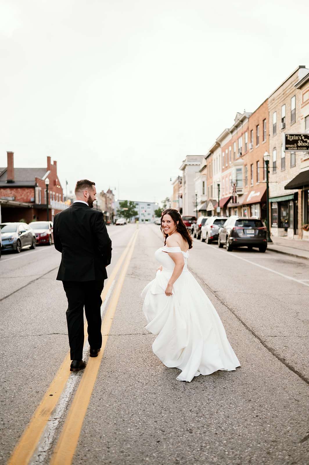 Bride and groom walk down the center of a small-town street, with the bride looking back and smiling playfully.