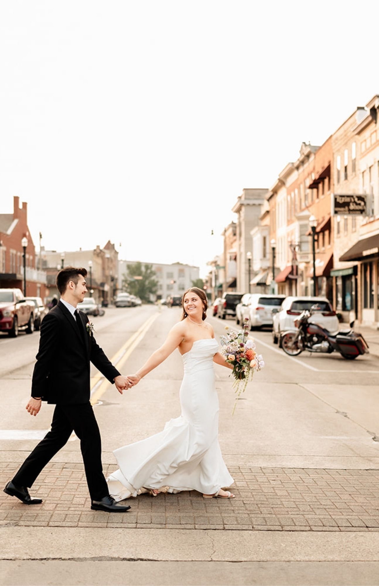 Just-married couple laughing as they walk across a downtown Burlington crosswalk, surrounded by historic buildings and parked cars.