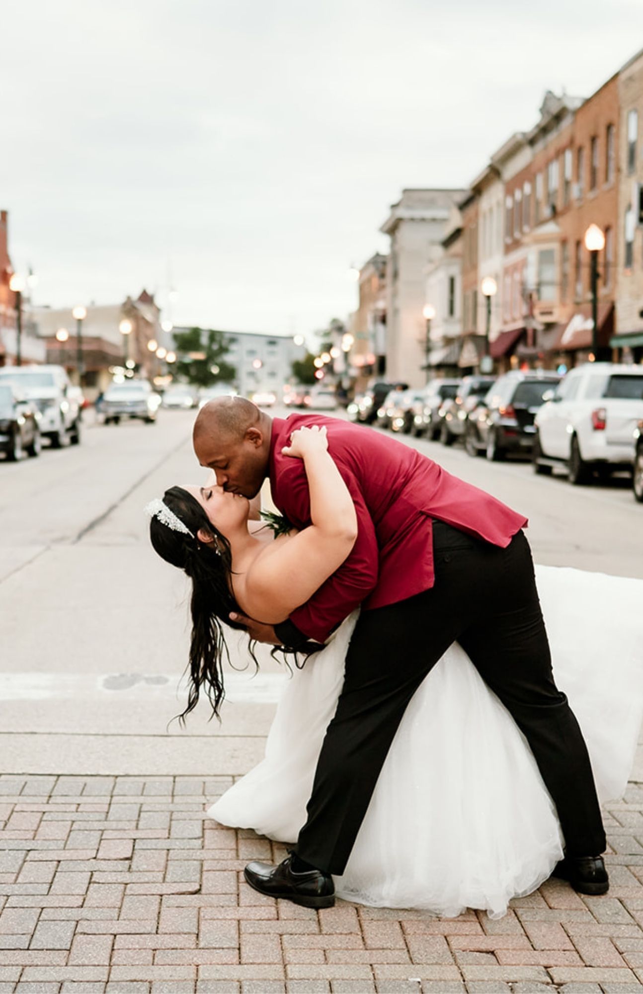 Bride and groom walking across the street in downtown Burlington, Wisconsin, with historic brick architecture and streetlights in the background.