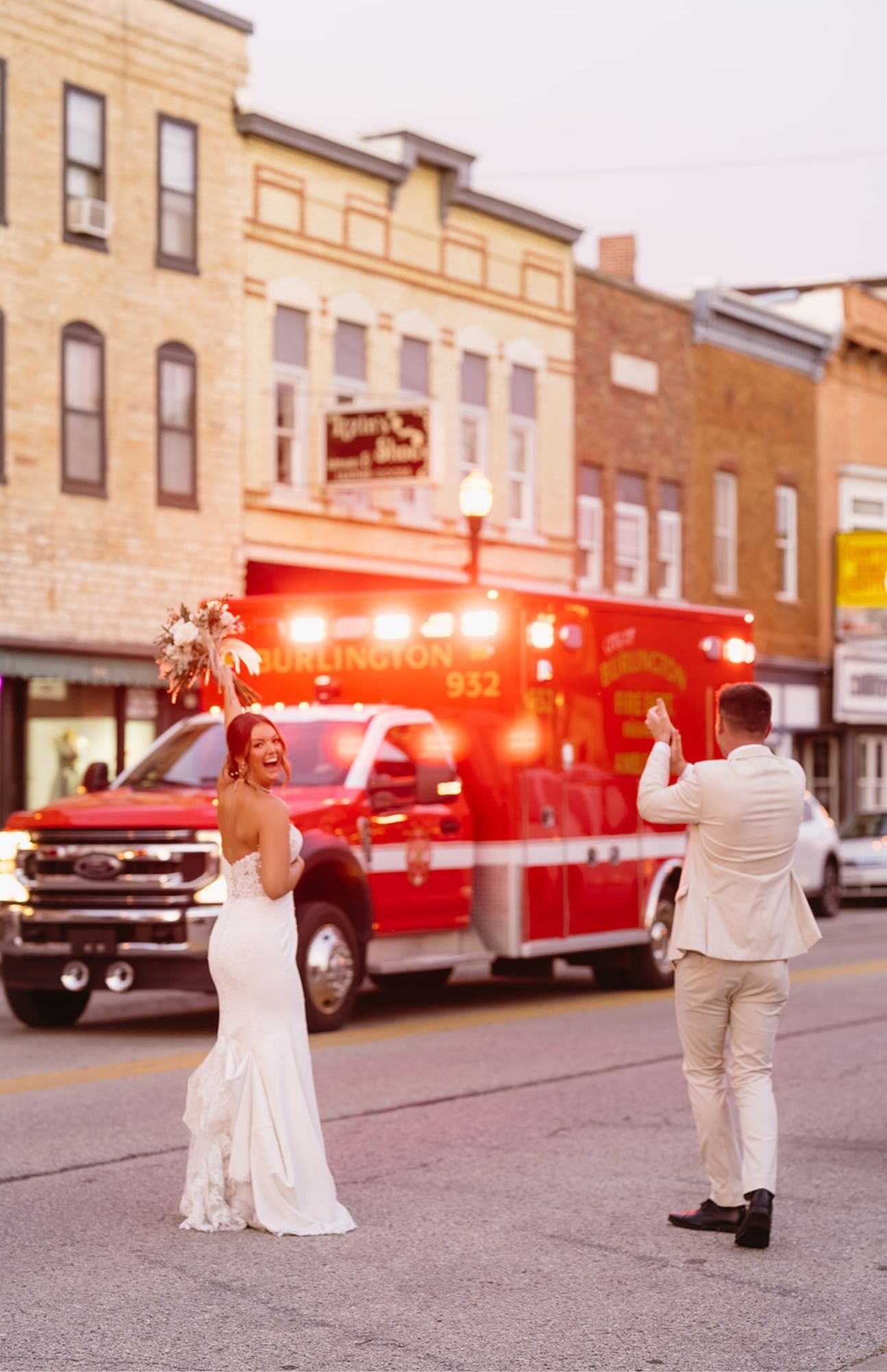 Wedding couple walking hand in hand past boutique shops in downtown Burlington, Wisconsin, cheering as an ambulance flashes its sirens to celebrate their marriage.