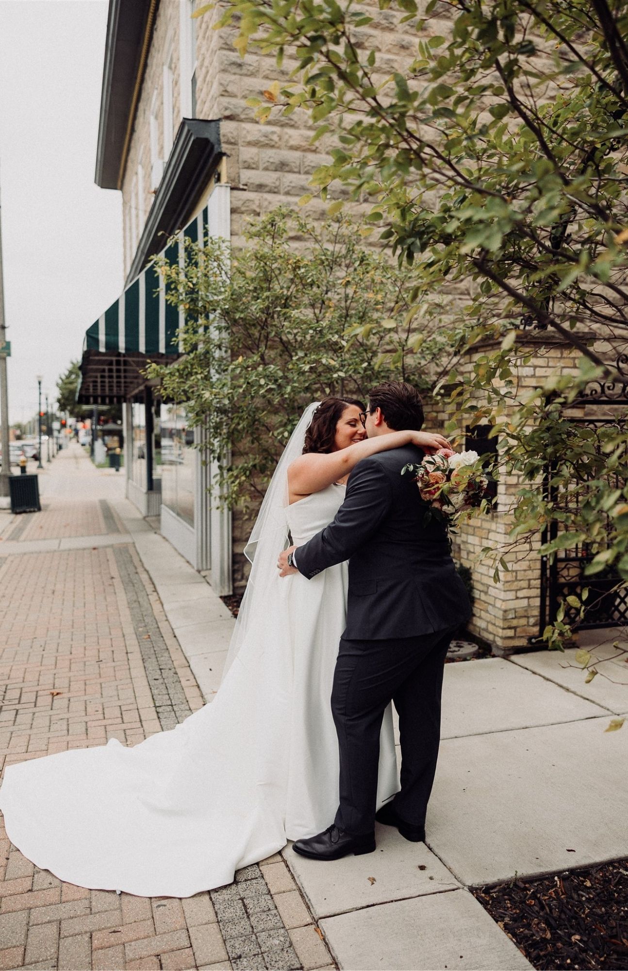 Just-married couple walking past a mural and brick buildings in downtown Burlington, Wisconsin, holding hands and smiling softly.