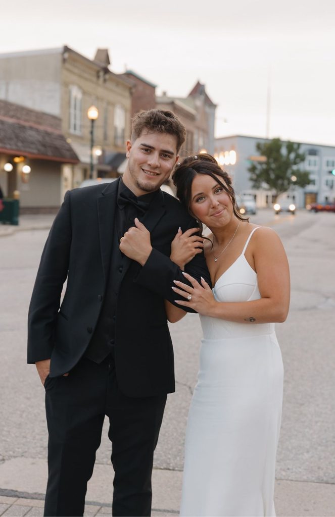 Bride and groom strolling past historic storefronts in downtown Burlington, Wisconsin, pausing to smile at each other in the soft afternoon light.