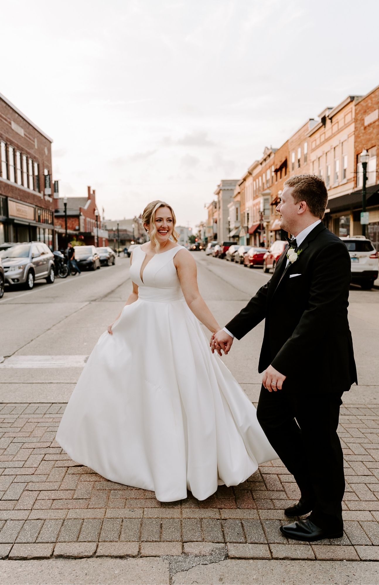 Newlyweds walking hand in hand past a brick wall and string lights in downtown Burlington, Wisconsin, during golden hour.